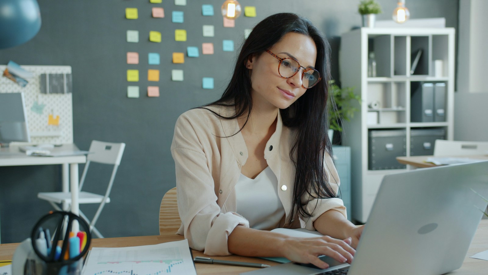 Young woman in glasses typing on a laptop at an office desk, with a wall of colourful sticky notes behind her