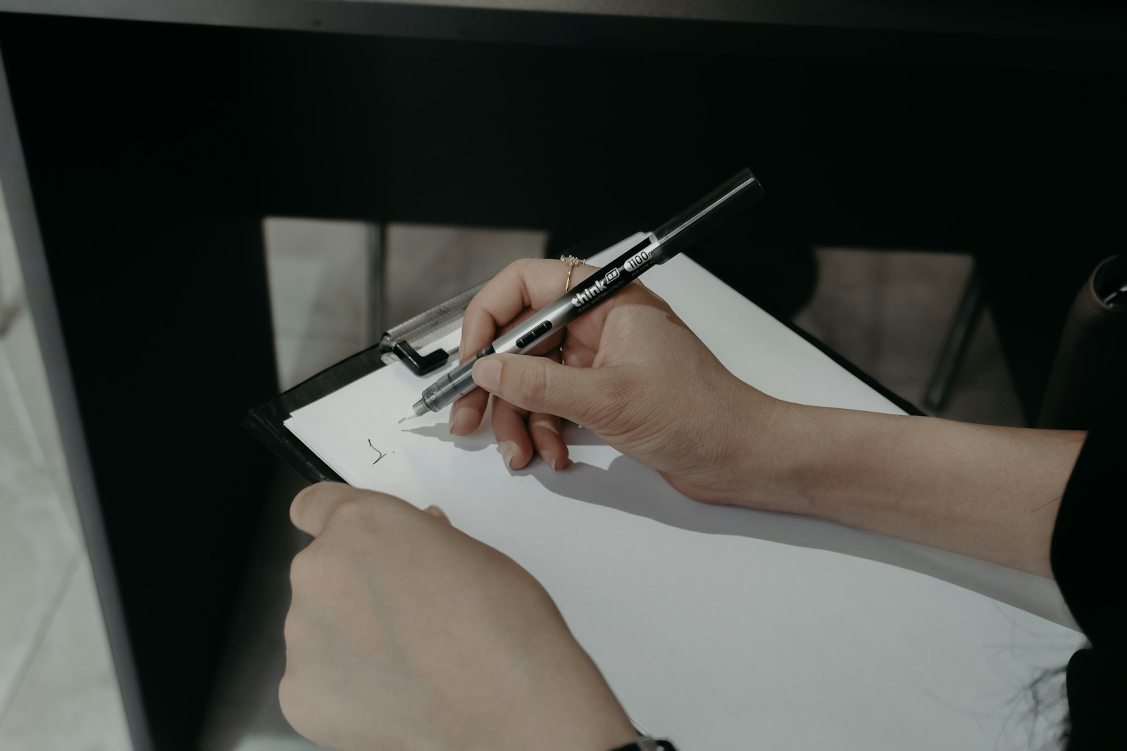 Two hands start a checklist, writing the number one on a blank sheet clipped to a black clipboard