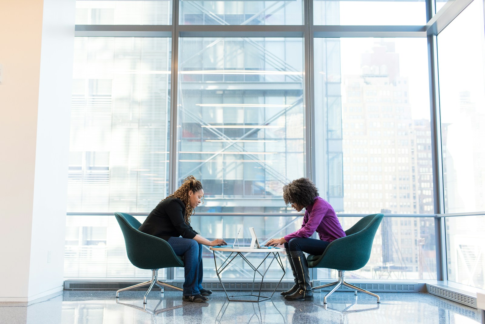 Two women type on laptops across a small round table in teal armchairs beside a tall office window