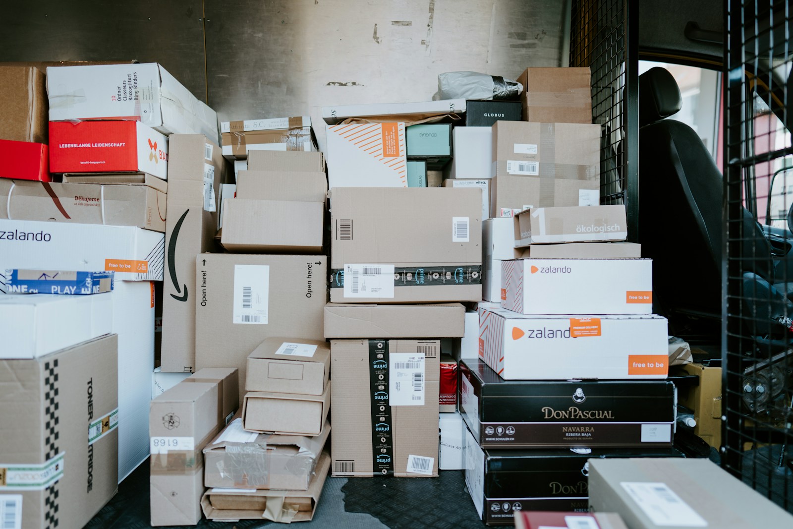 Parcels and shipping boxes with barcode labels stacked high in a delivery van's cargo area behind the driver's seat