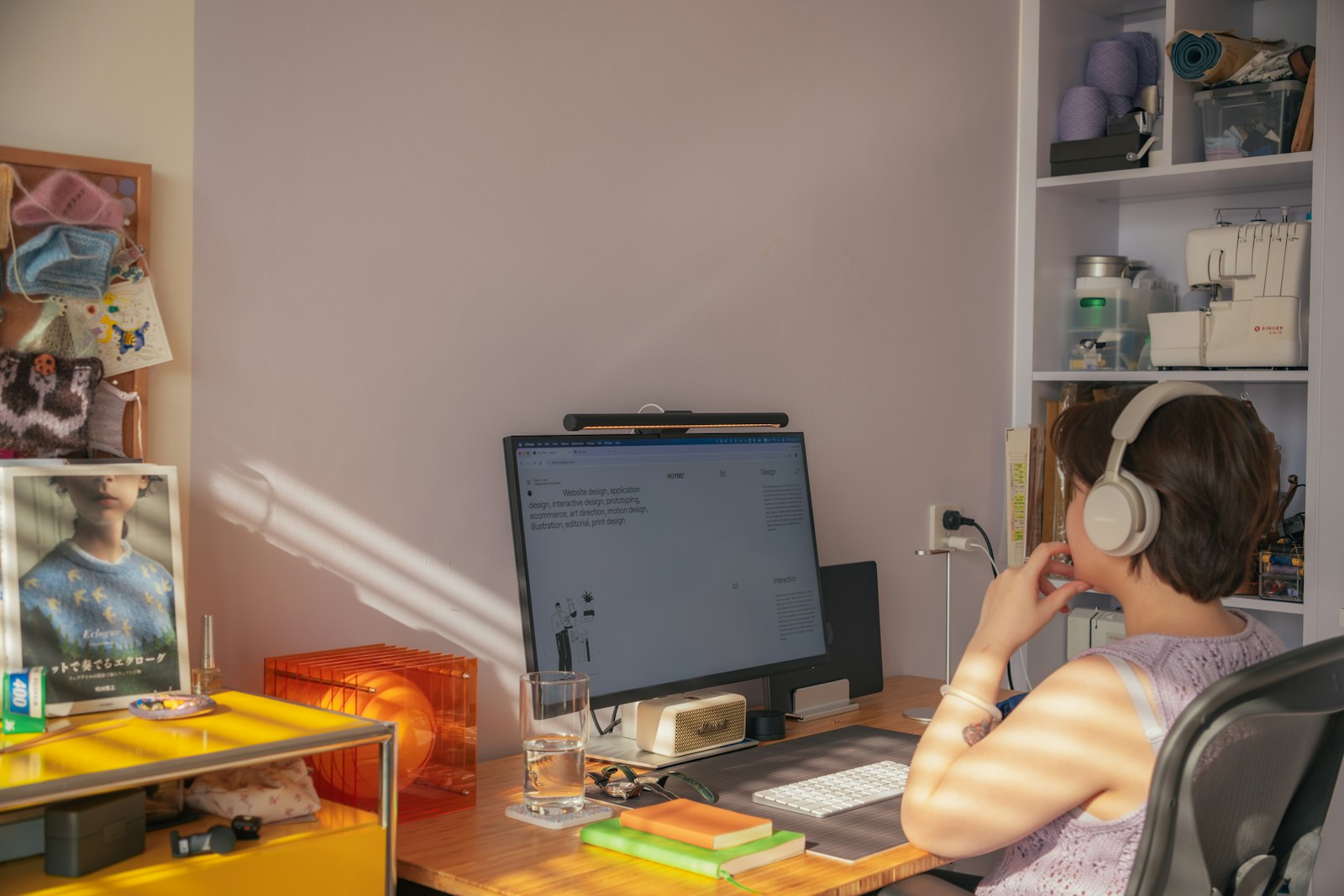 A woman in headphones works at a monitor on a sunlit desk beside shelves holding a sewing machine and yarn