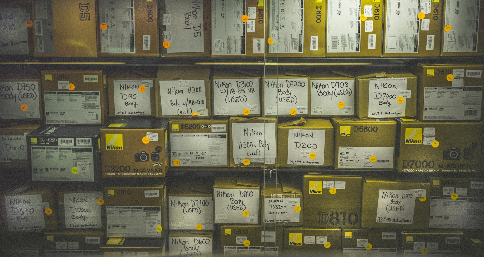 Shelves of boxed used Nikon cameras with handwritten model labels and small yellow stickers marked WEB