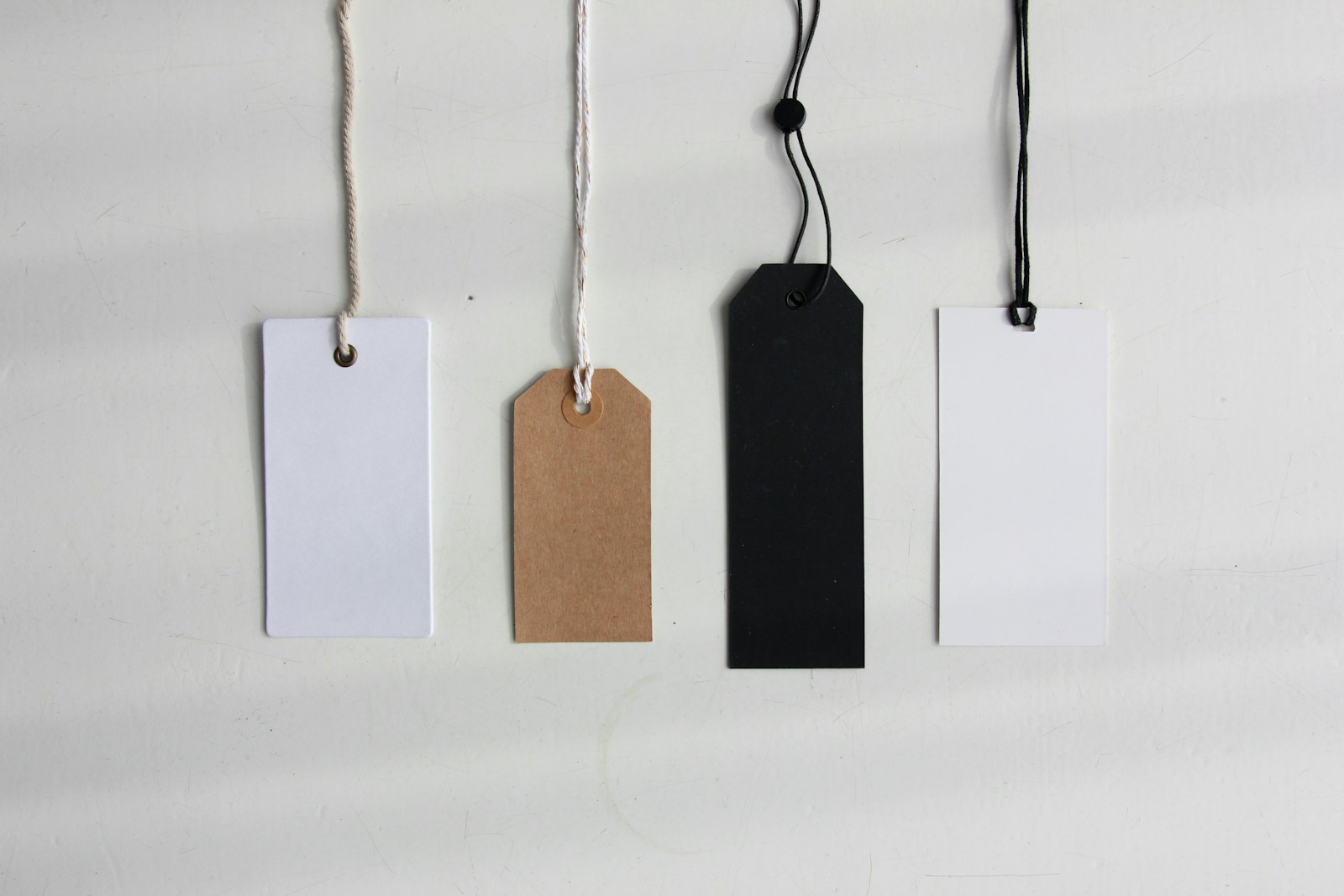 Four blank price tags in white, kraft brown, black and white hanging on strings against a pale wall