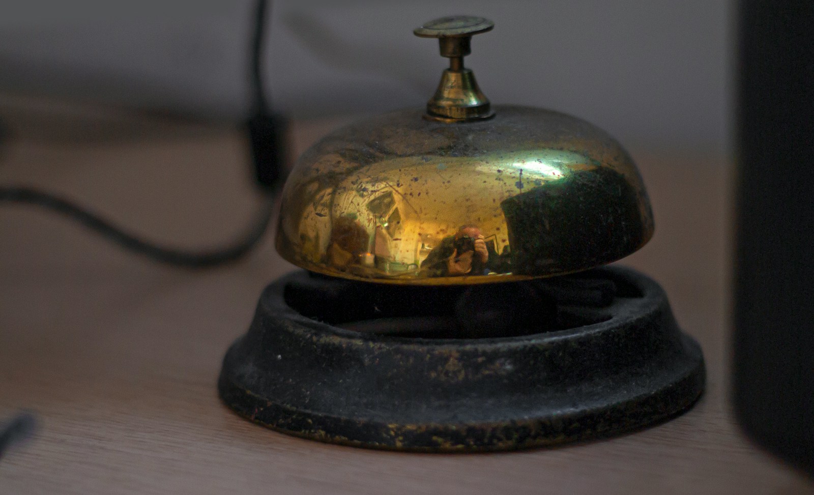 A worn brass service bell on a wooden desk, its polished dome reflecting the photographer and the room
