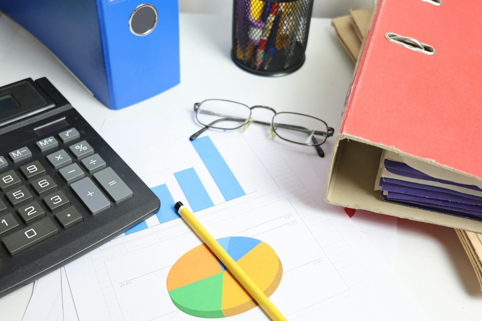 A black desk calculator, printed bar and pie charts, a yellow pencil and reading glasses on a desk between blue and red binders