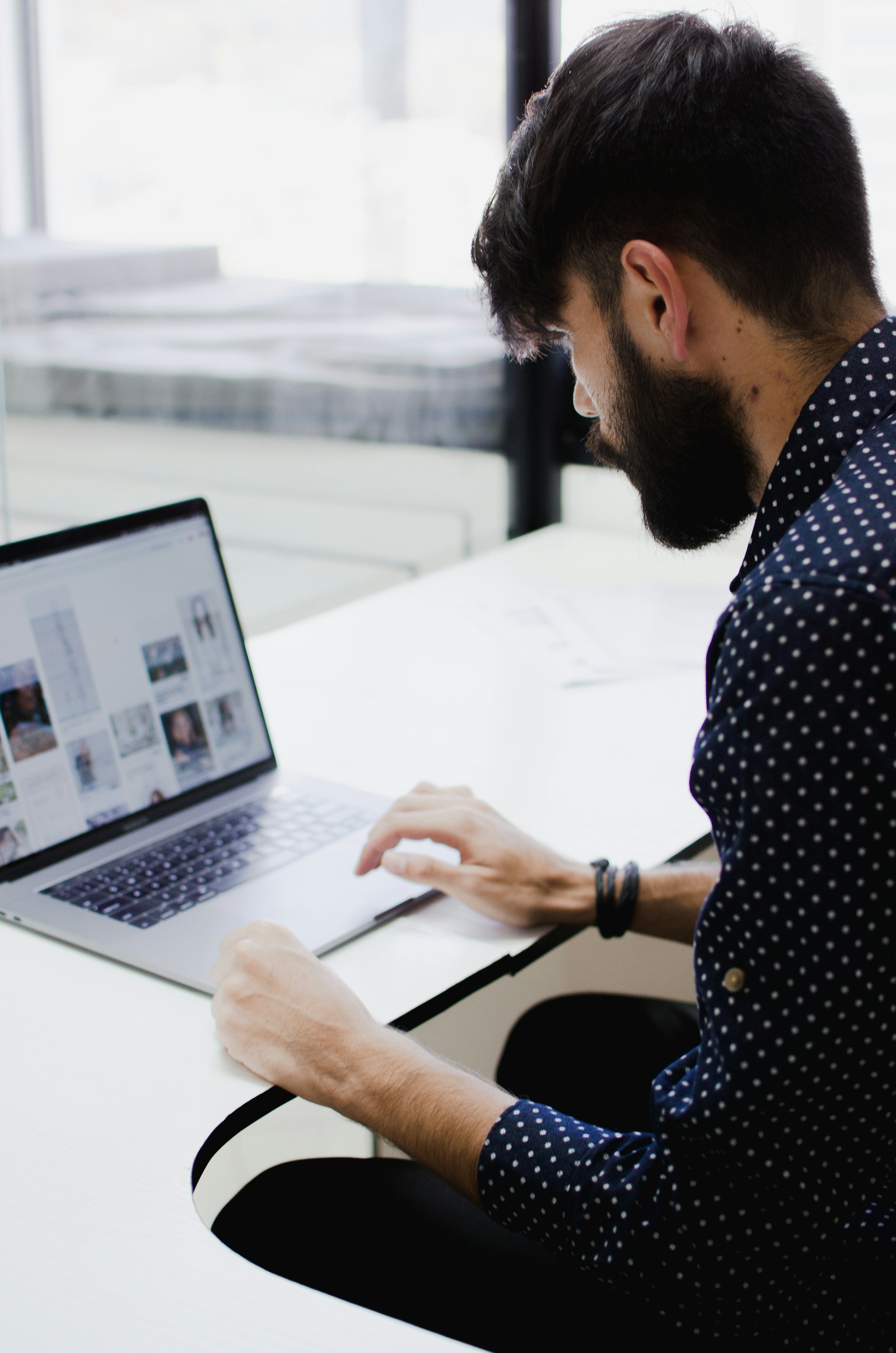 A bearded man in a polka-dot shirt works at a laptop on a white desk beside a bright window