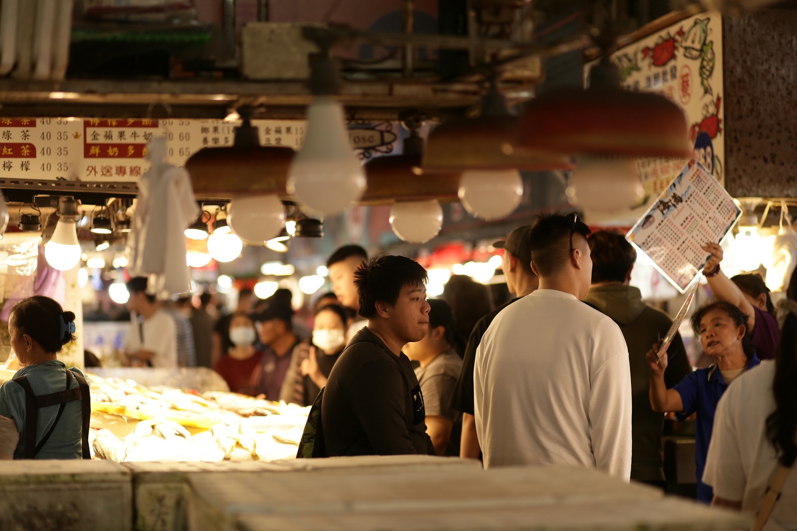 Shoppers crowd a busy indoor market under hanging lamps while a vendor holds up a printed menu