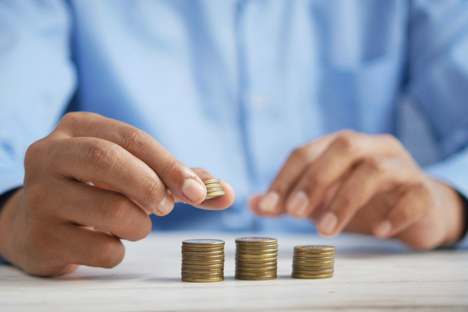 A person in a light blue shirt places a coin on one of three small stacks of coins on a white table