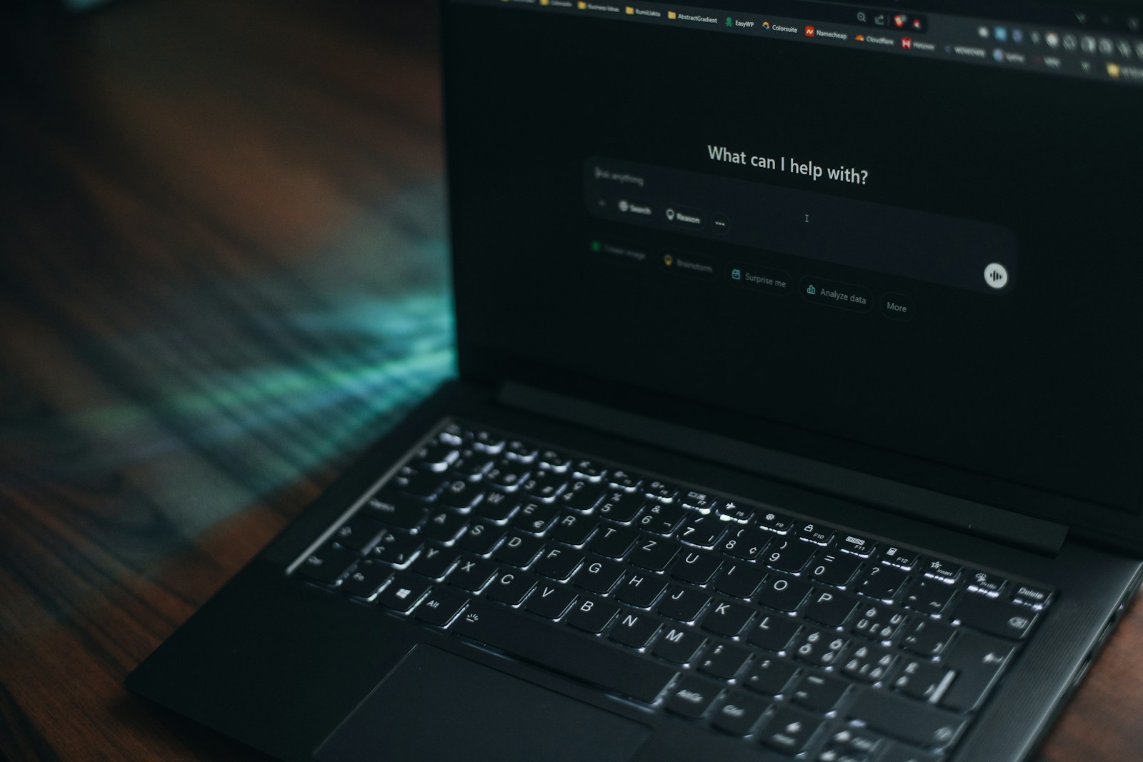 A dark laptop on a wooden table showing an AI assistant's empty prompt box under the heading What can I help with?
