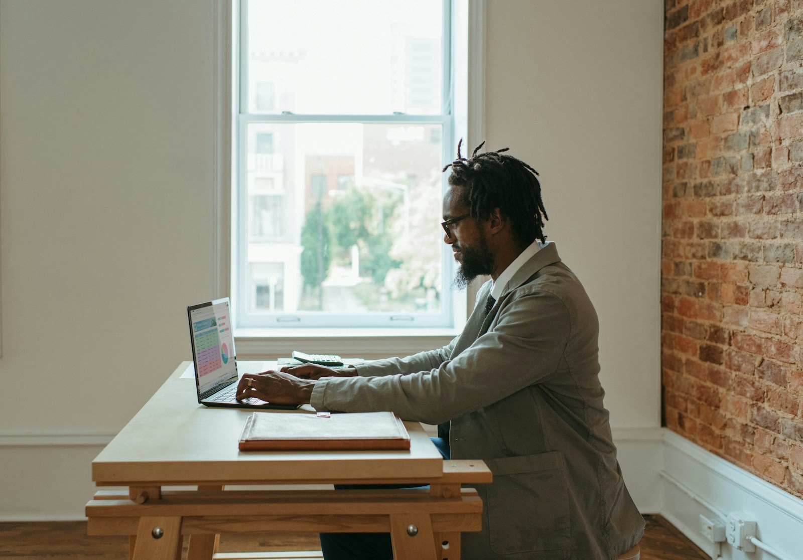 A man in glasses and a grey jacket types on a laptop at a wooden desk beside a tall window