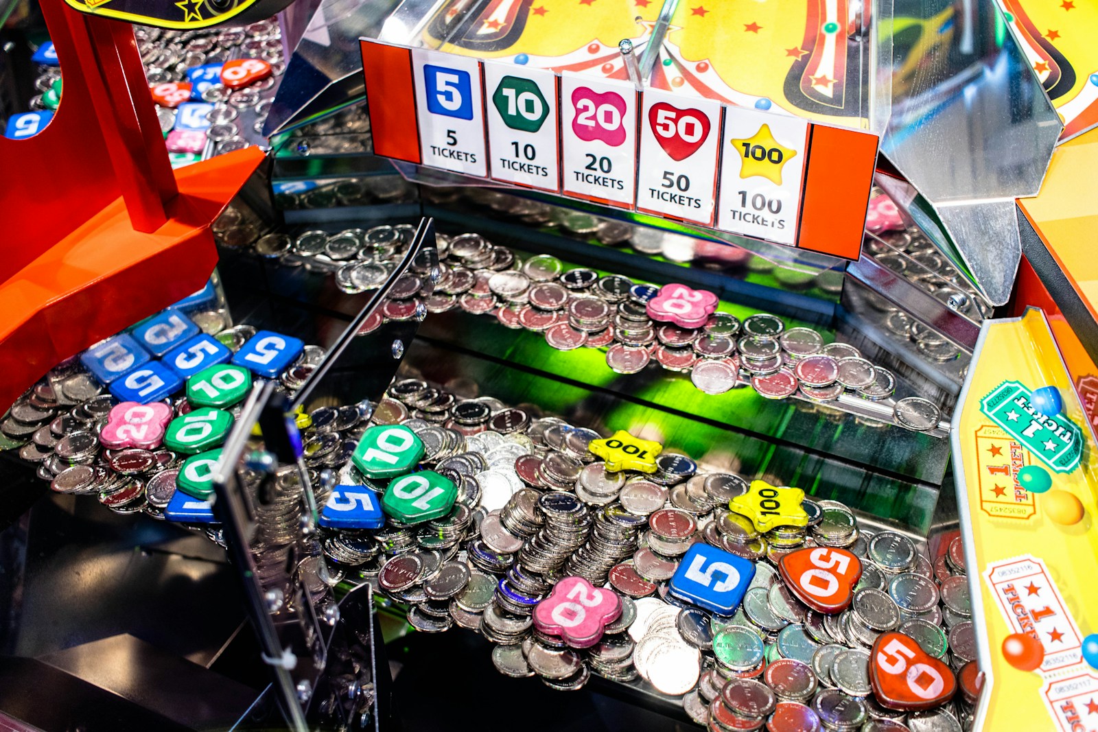 Colourful arcade tokens marked 5, 10, 20, 50 and 100 lie among silver coins inside a coin-pusher machine