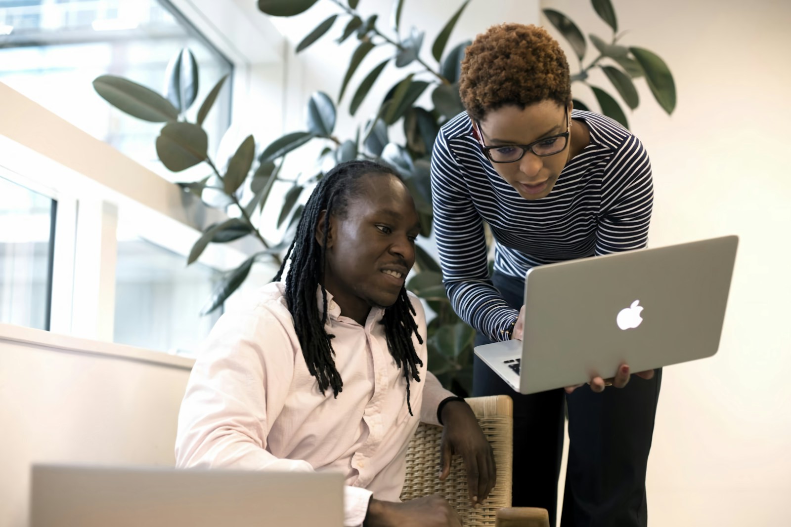 Two women lean over a laptop at a bright office desk, one of them holding the screen while they read it together