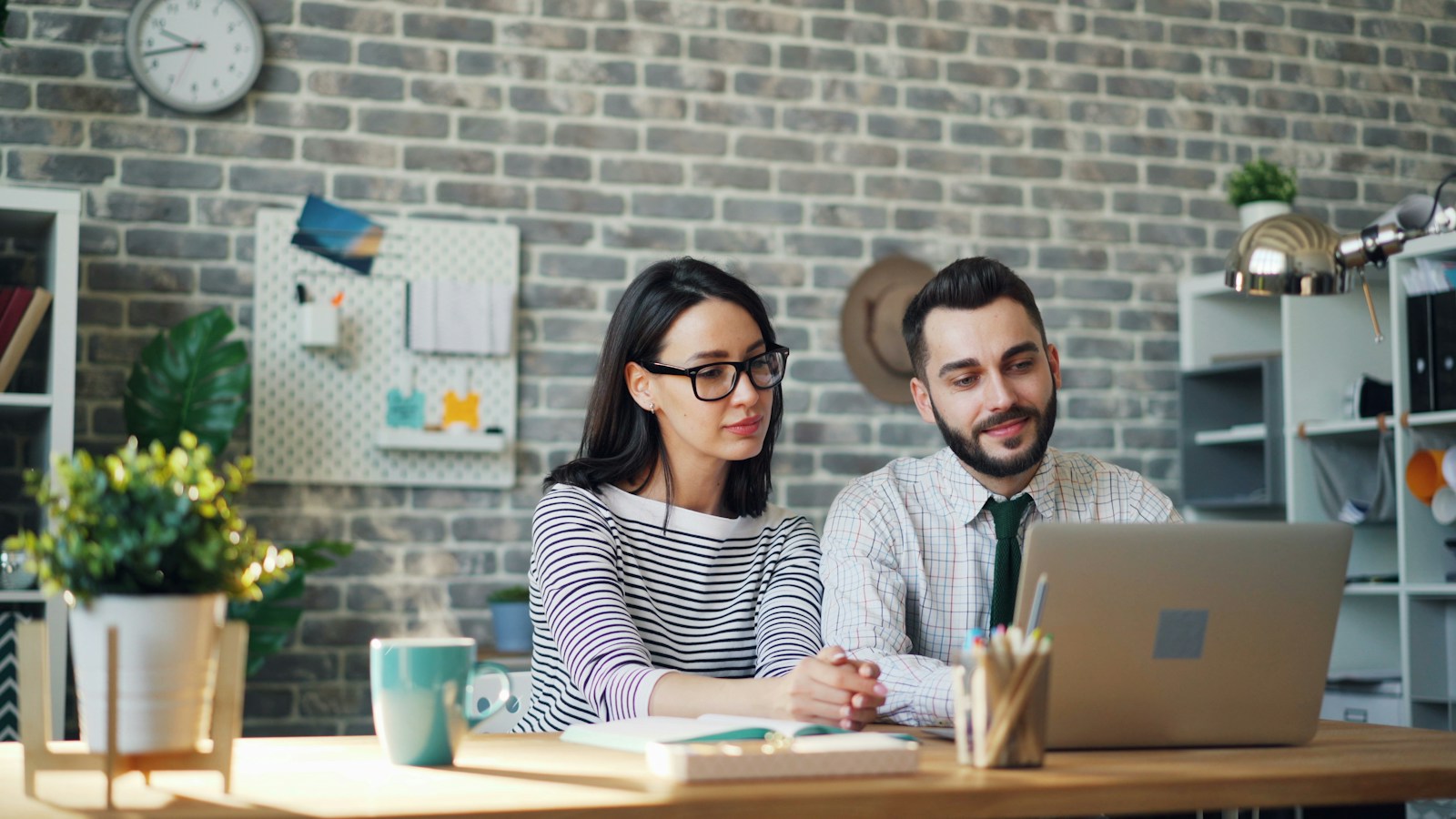 A woman in glasses and a man in a tie sit together at a wooden desk looking at one laptop in a brick-walled office