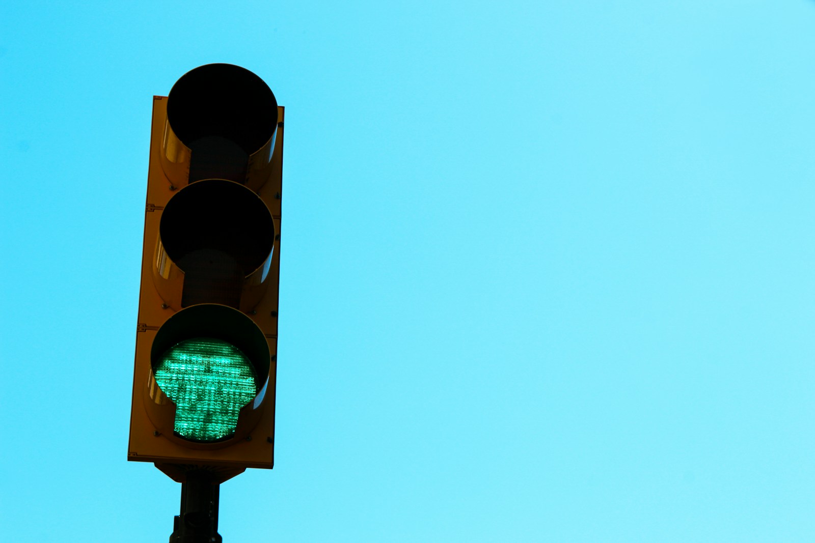A yellow traffic light against a clear blue sky with only its green signal lit