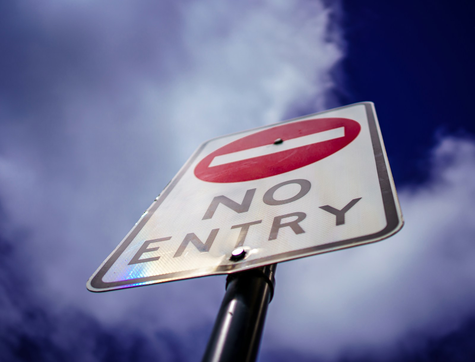 A white No Entry road sign with a red circle and white bar, seen from below against a deep blue sky and clouds