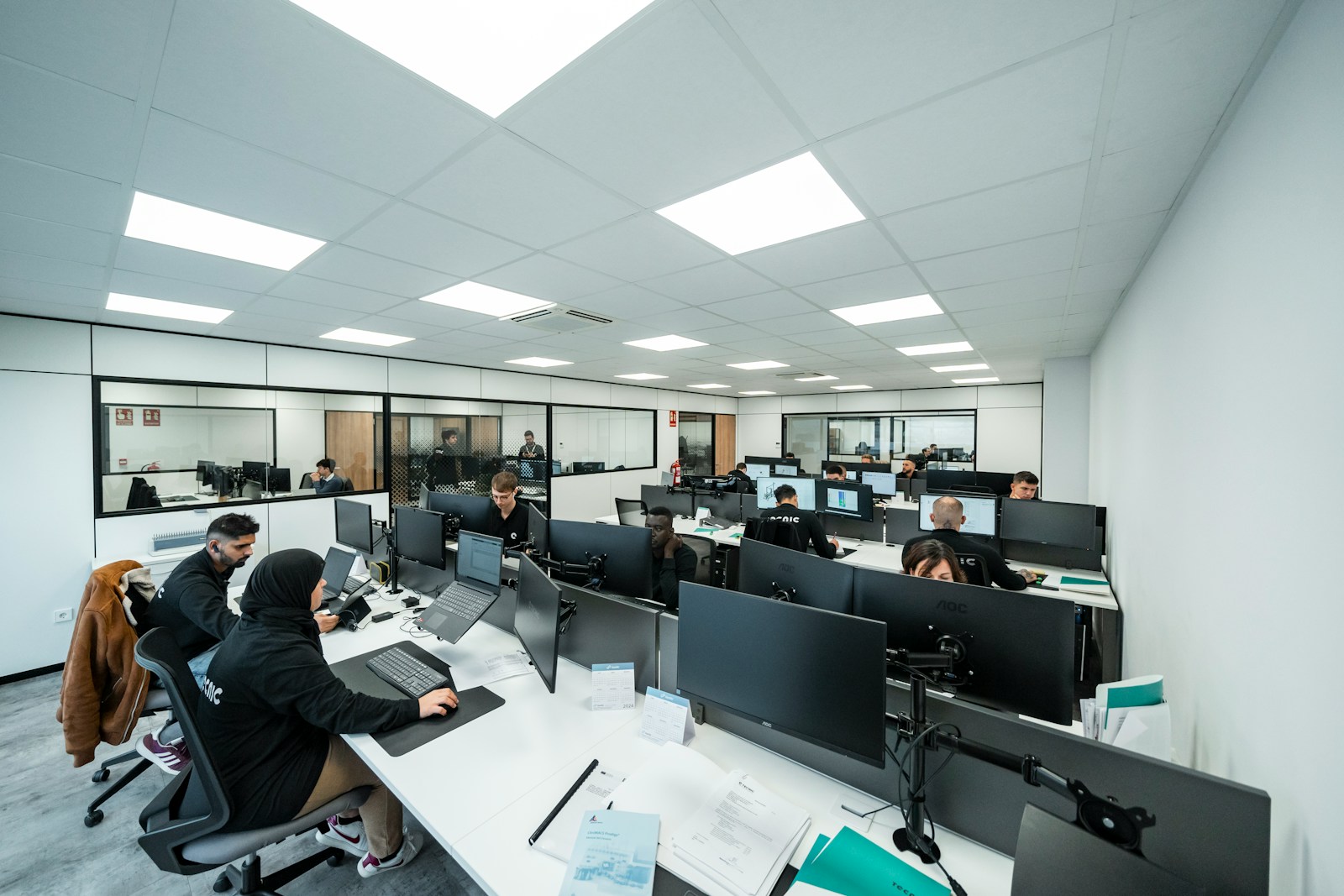 A large open-plan office where rows of staff work at desks with dual monitors, one man wearing a wireless earpiece