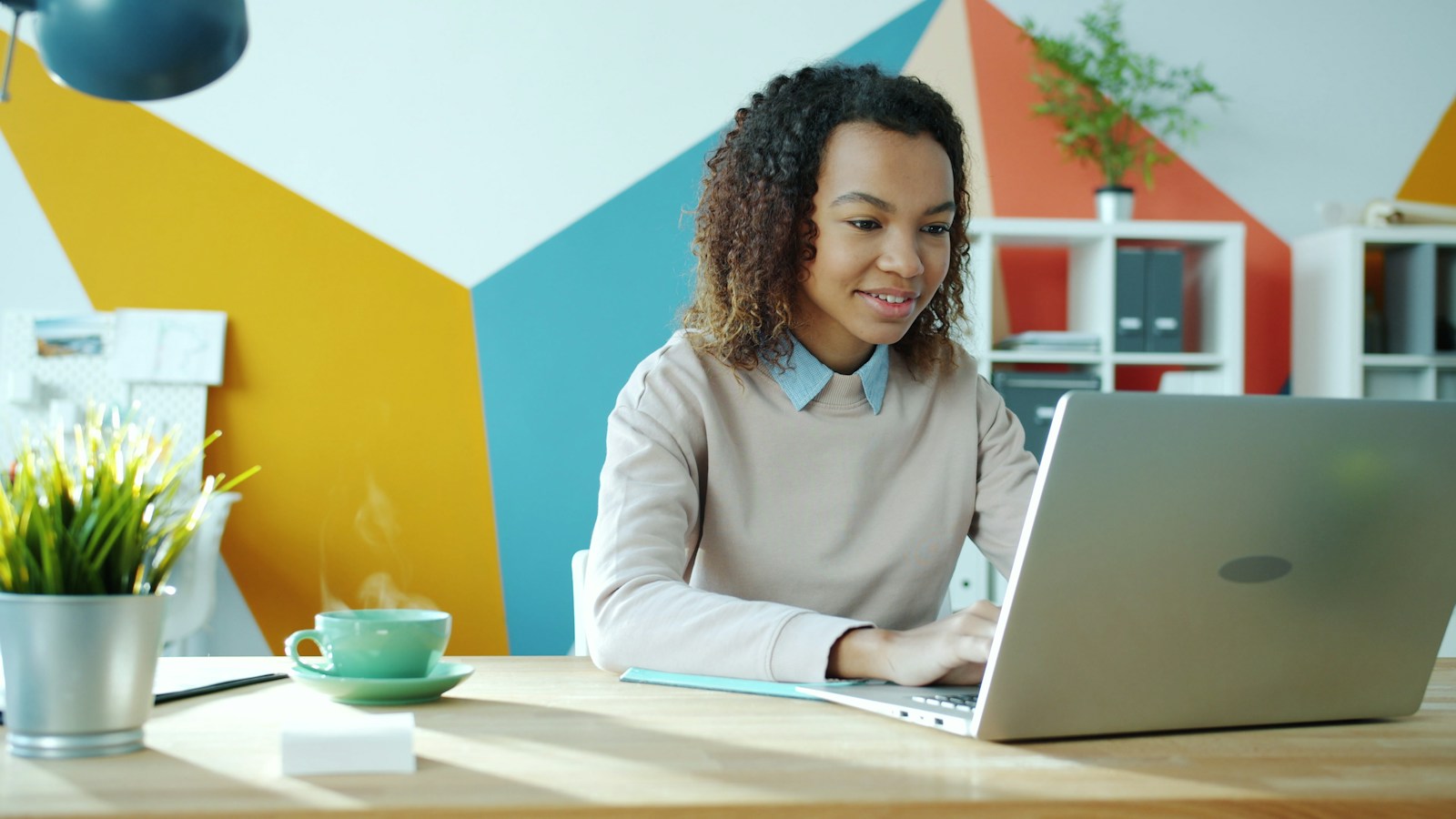 Smiling woman typing on a laptop at a wooden desk in a brightly painted office, with a steaming cup of tea beside her