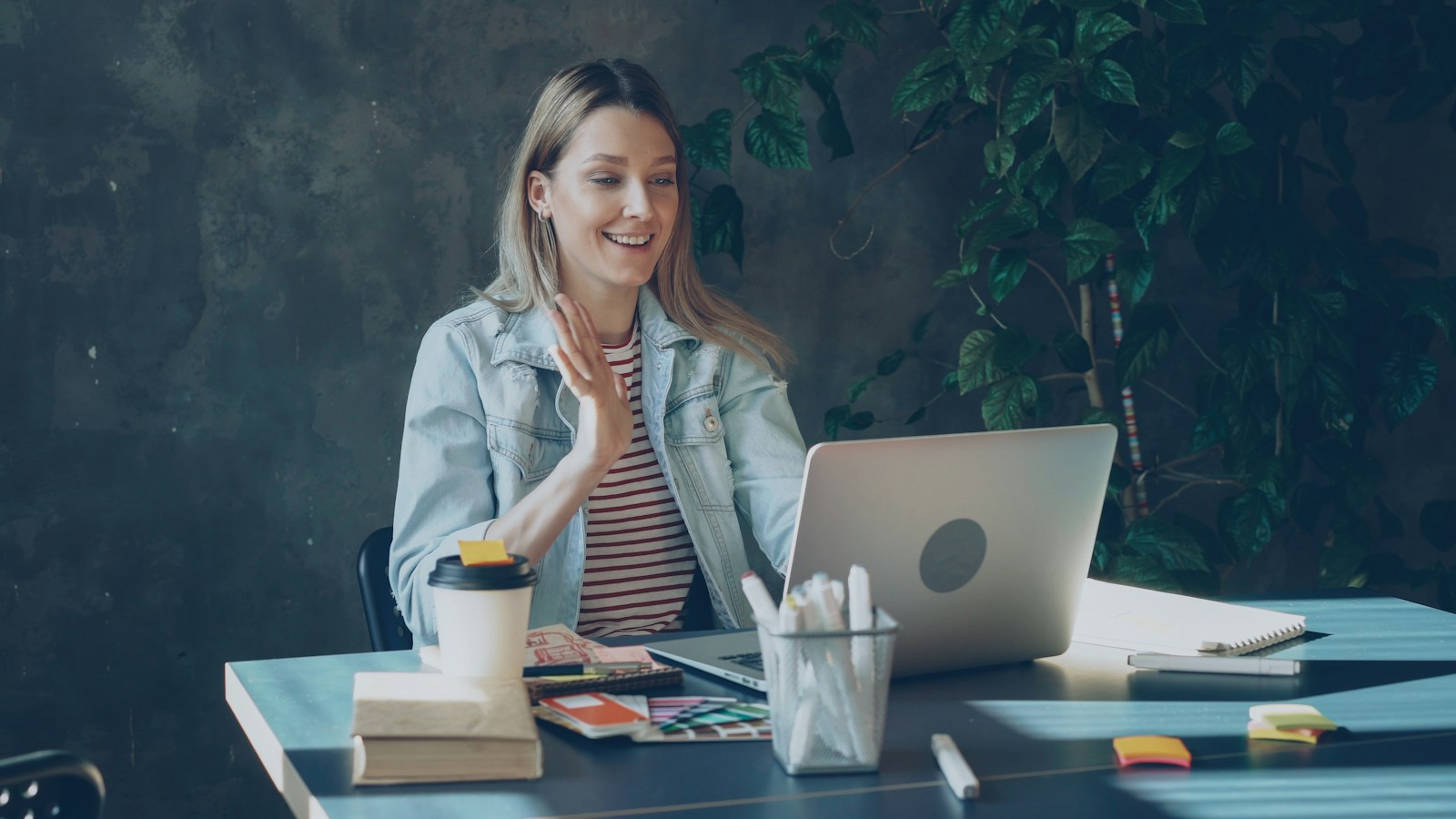 A smiling woman in a denim jacket waves at her laptop during a video call, at a desk with a coffee cup, pens and notebooks