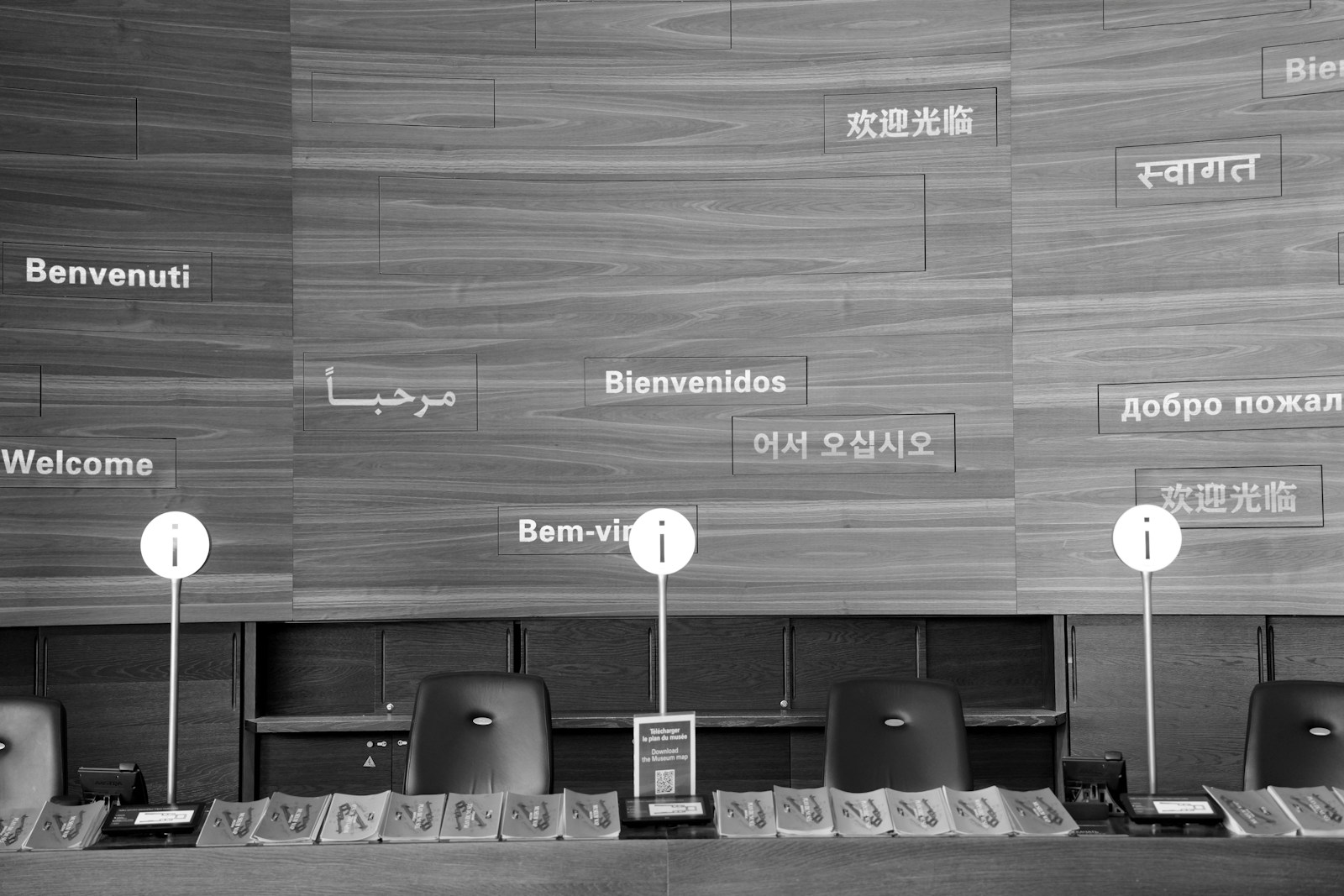 Black and white photo of a museum information desk with empty chairs and welcome greetings in many languages mounted on a wooden wall