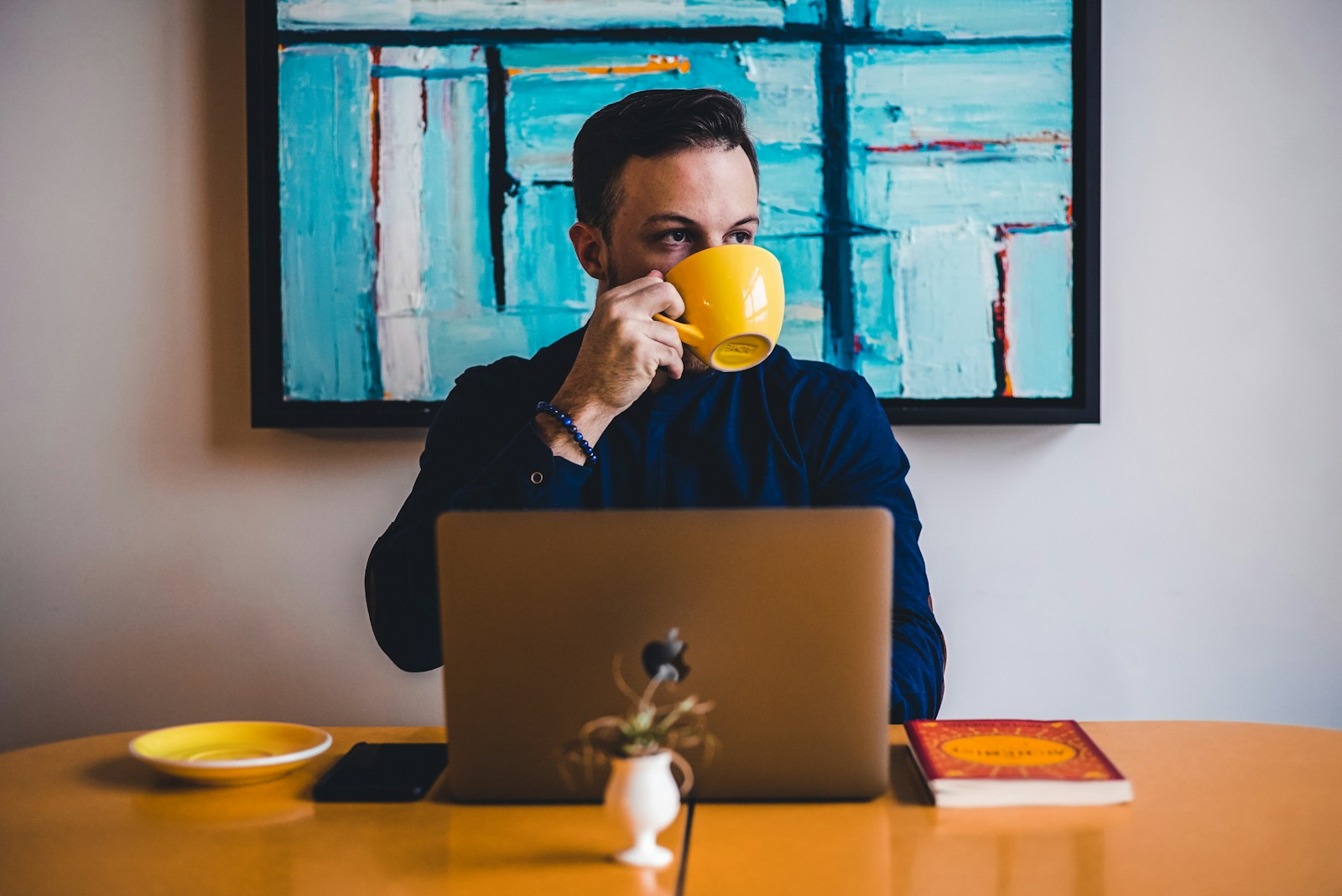 A man in a dark blue shirt sips from a yellow cup behind an open laptop at an orange table