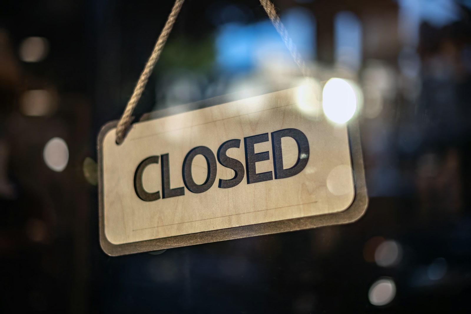 A wooden CLOSED sign hangs on a rope behind a shop's glass door, with blurred evening lights in the background
