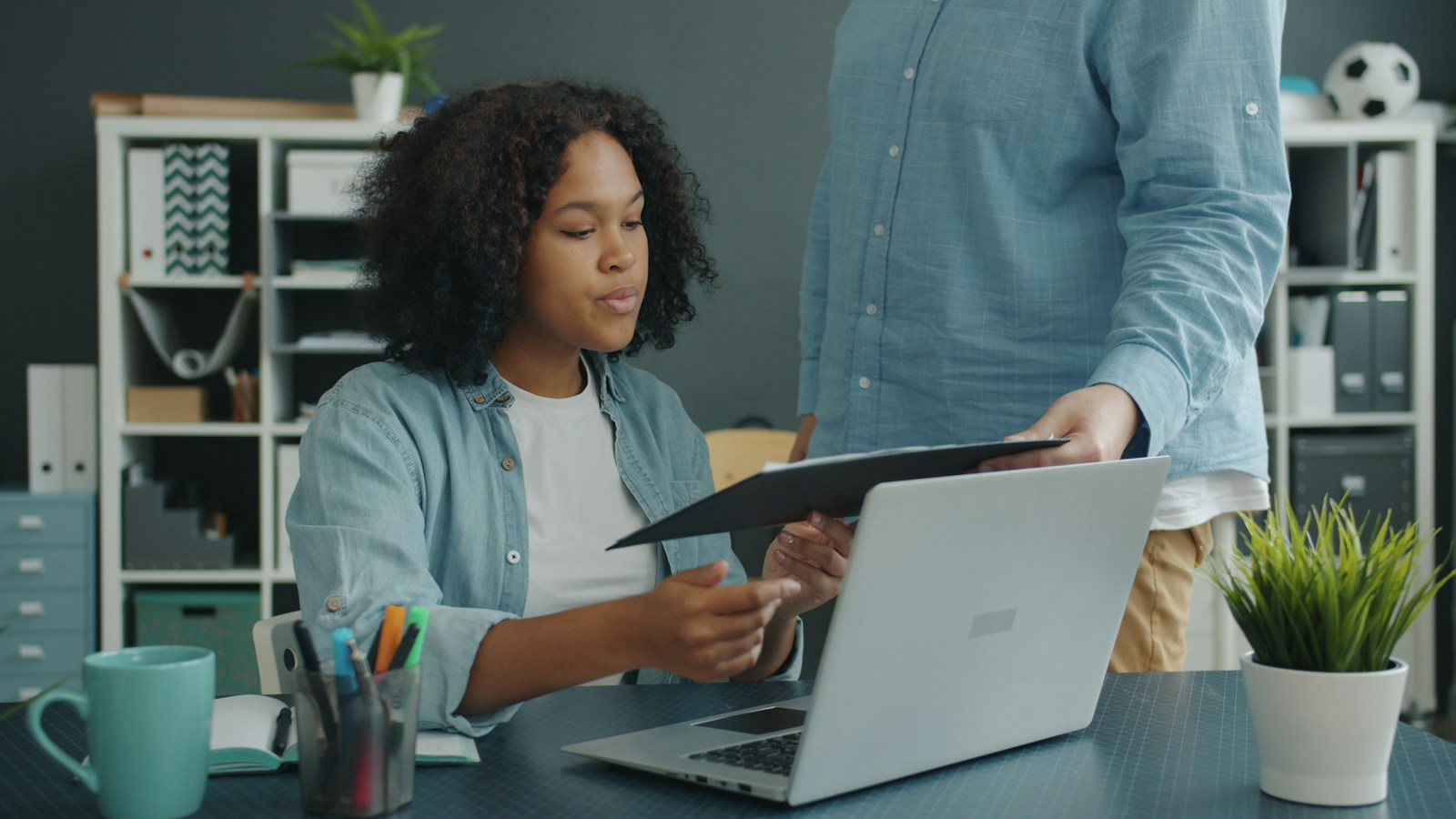 A woman at a laptop takes a dark folder from a colleague in a blue shirt standing beside her desk