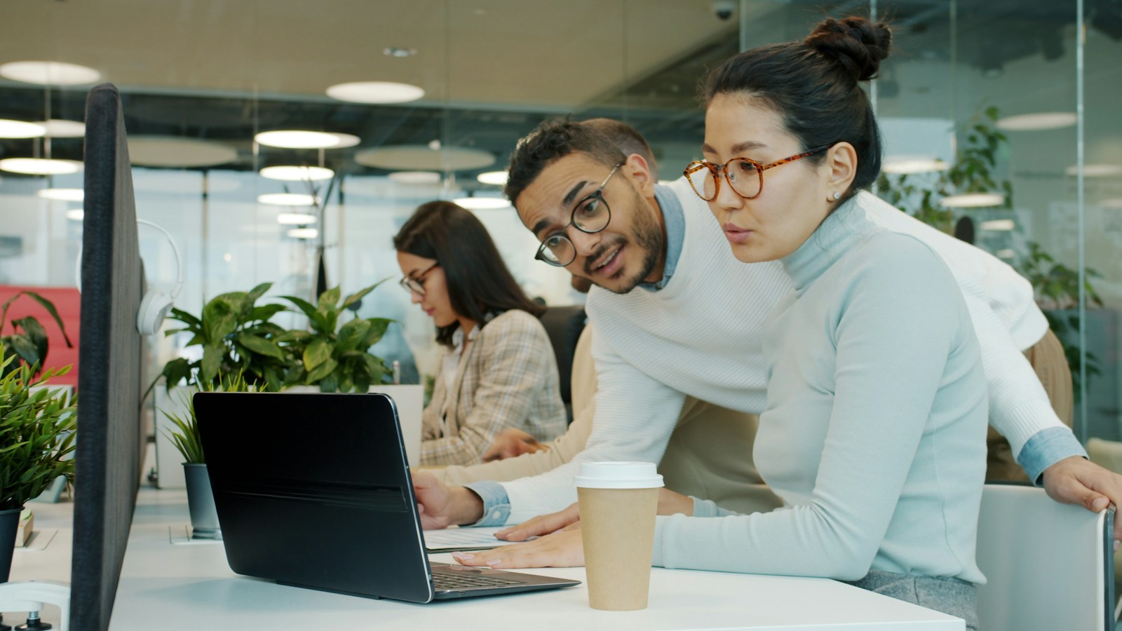 Two colleagues in glasses lean over a laptop together at a shared desk in an open-plan office