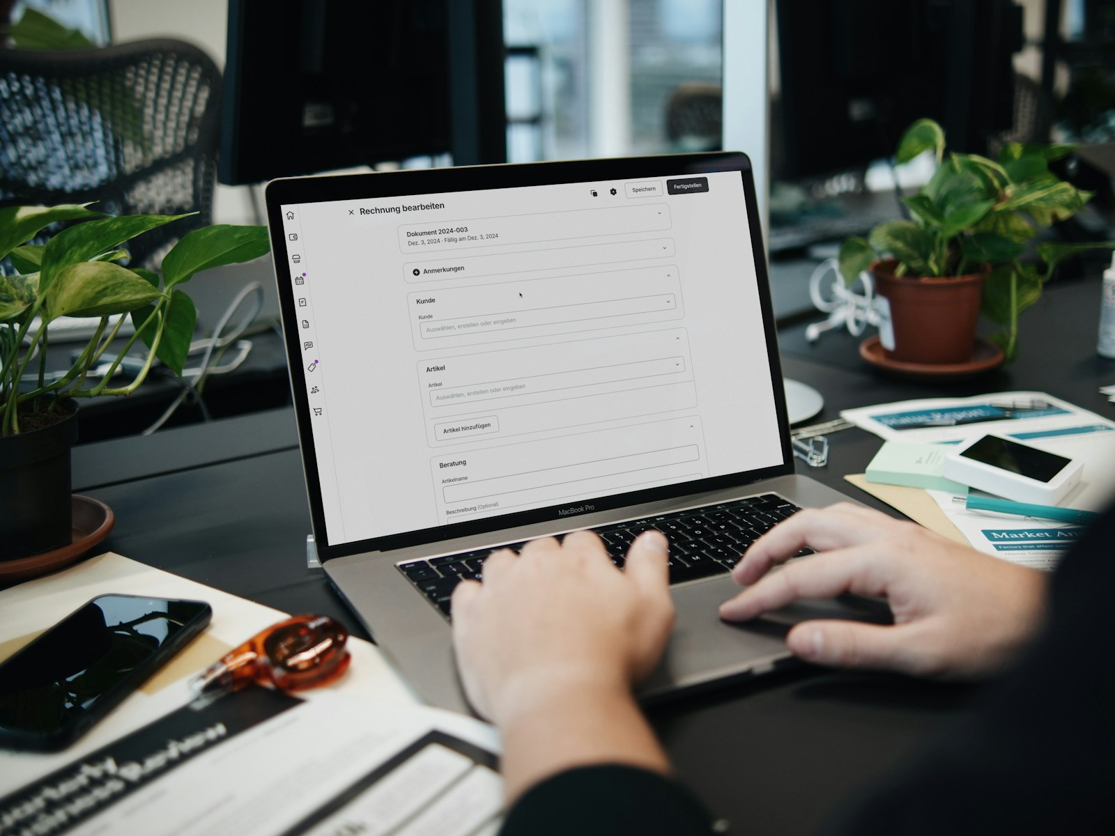 Hands typing on a laptop that shows an invoice-editing form, on a desk with potted plants, a smartphone and printed papers