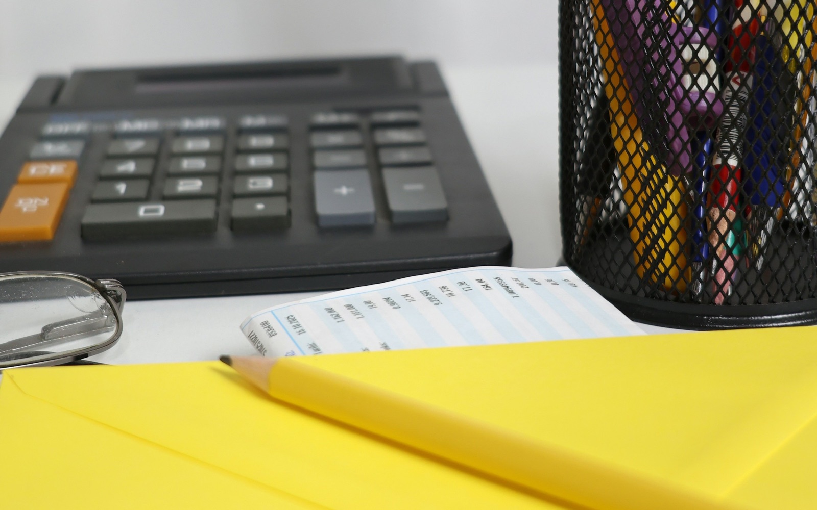 A black desk calculator, reading glasses and a printed slip of figures beside a yellow envelope and a pencil