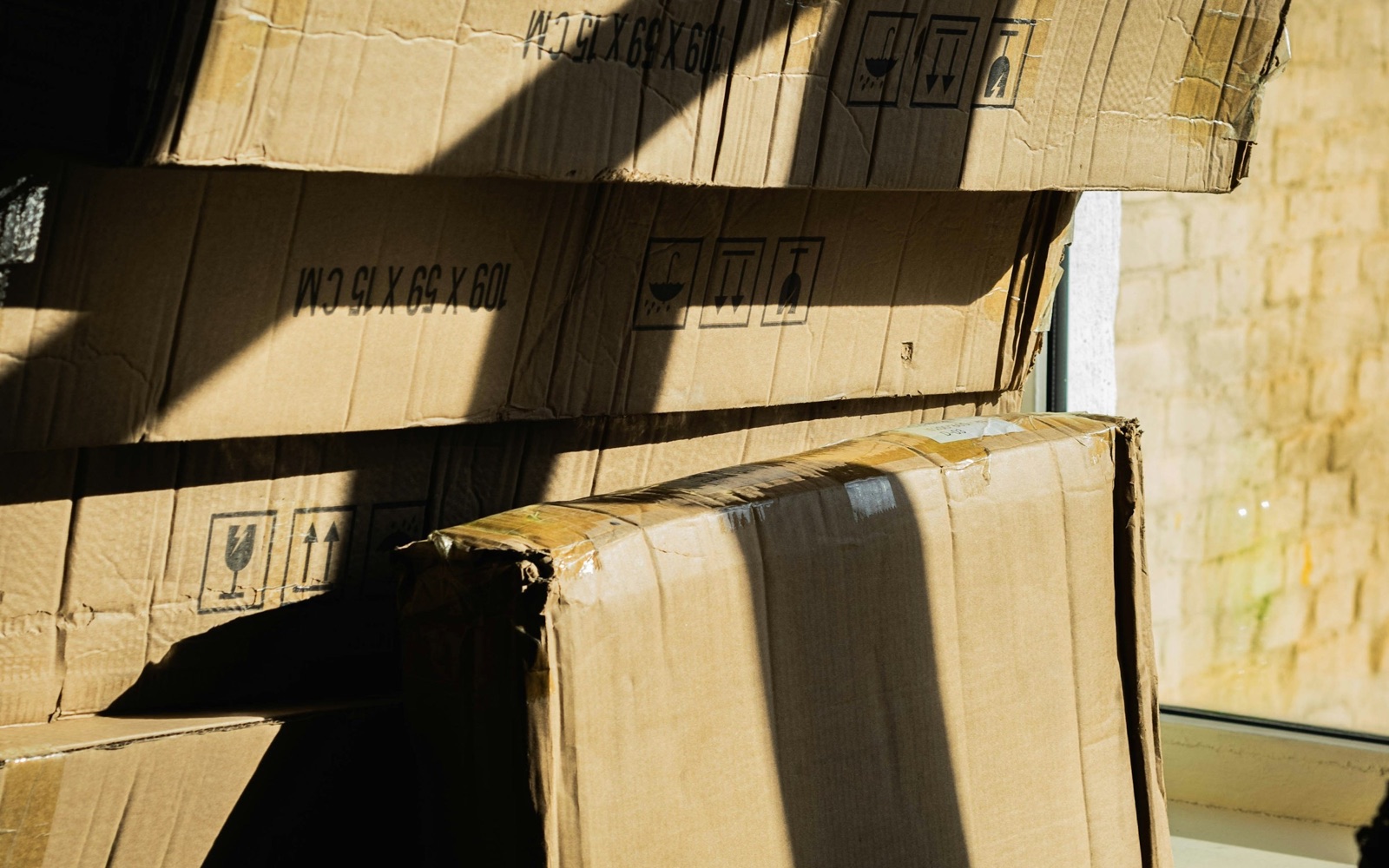Worn cardboard shipping cartons stacked by a sunny window, printed with their dimensions and handling symbols for fragile, upright and dry