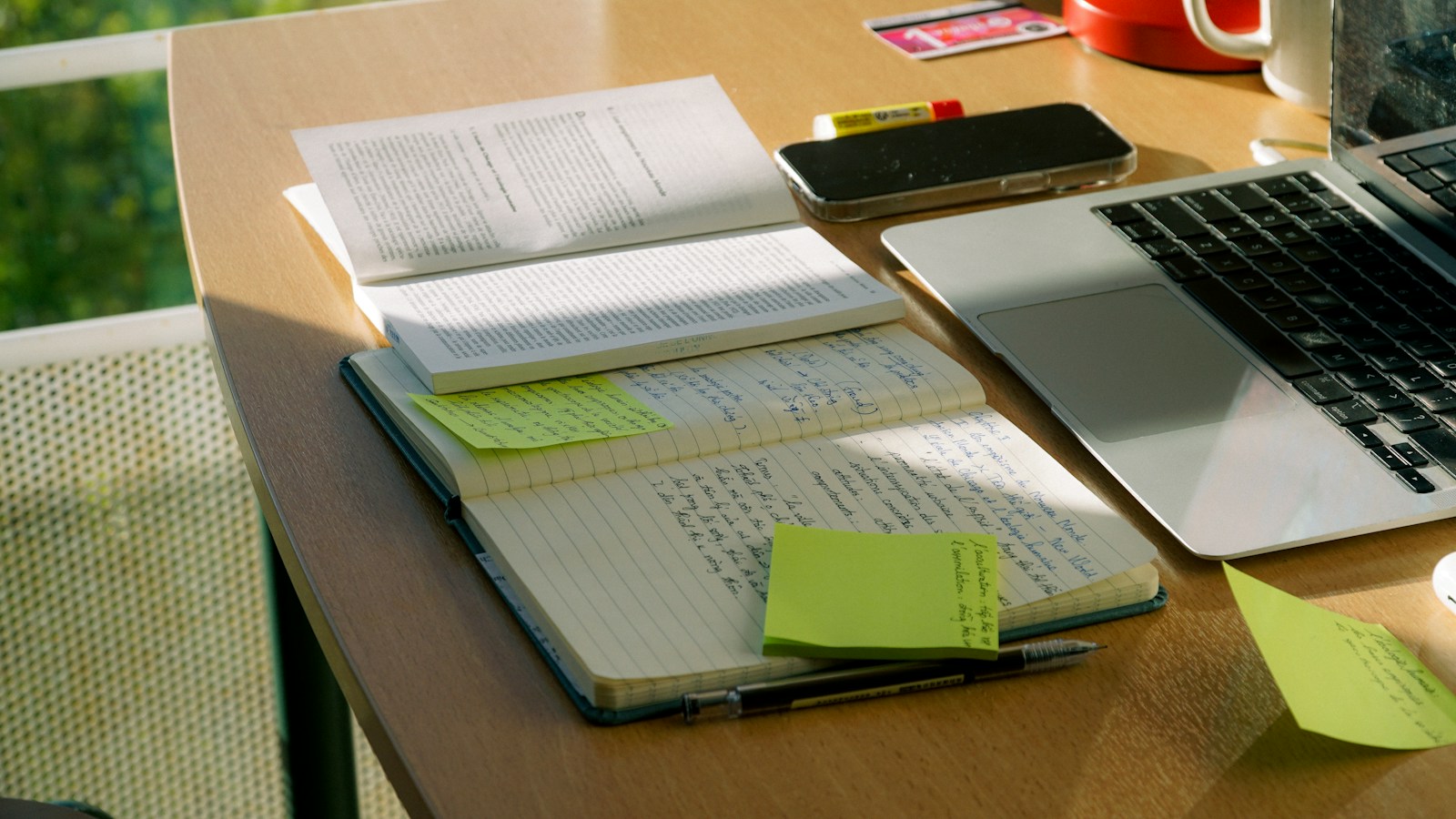 An open notebook with handwritten notes and green sticky notes lies beside a laptop keyboard on a sunlit desk