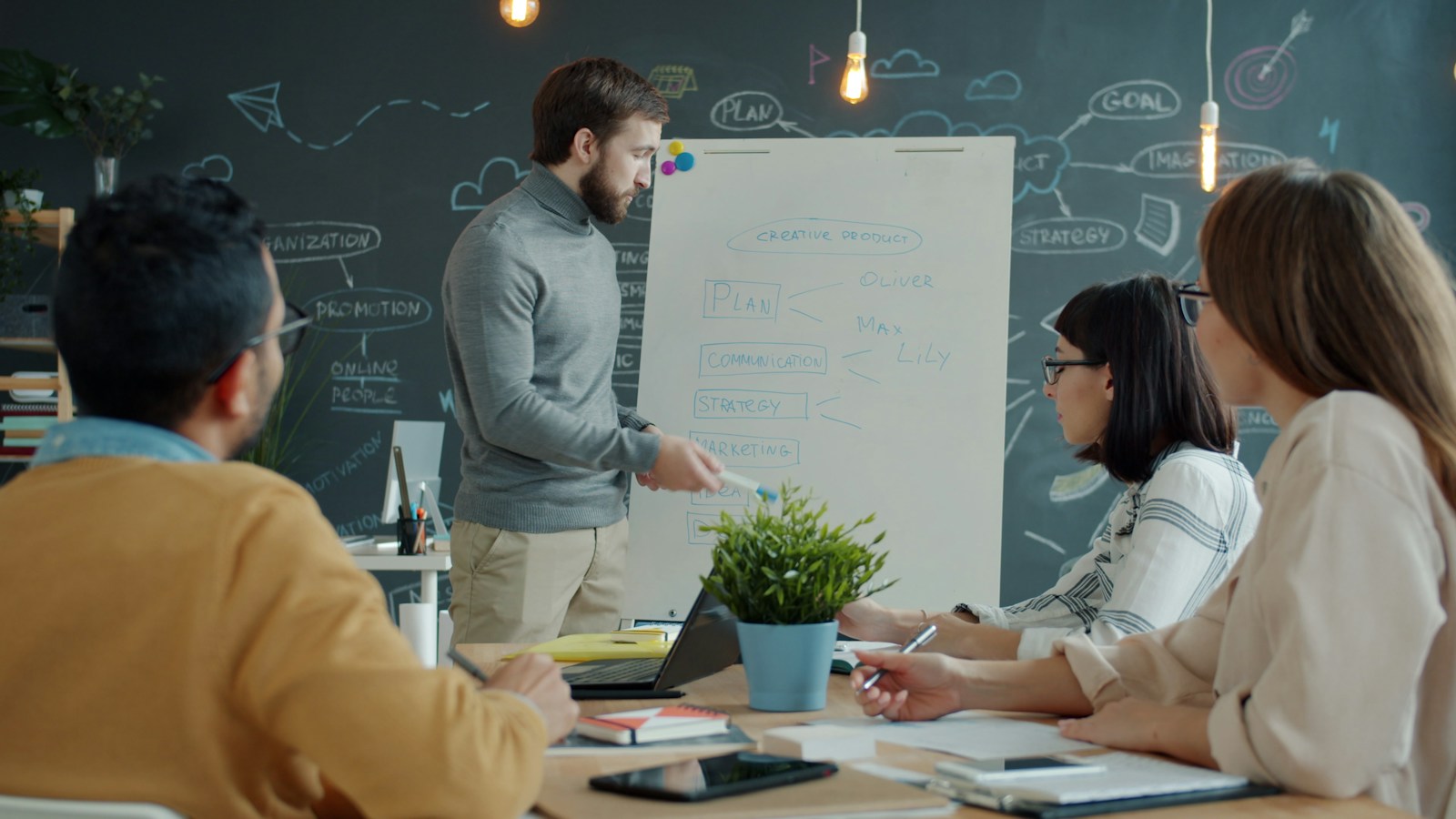 A man in a grey turtleneck presents a plan on a flipchart to three colleagues seated around a wooden table