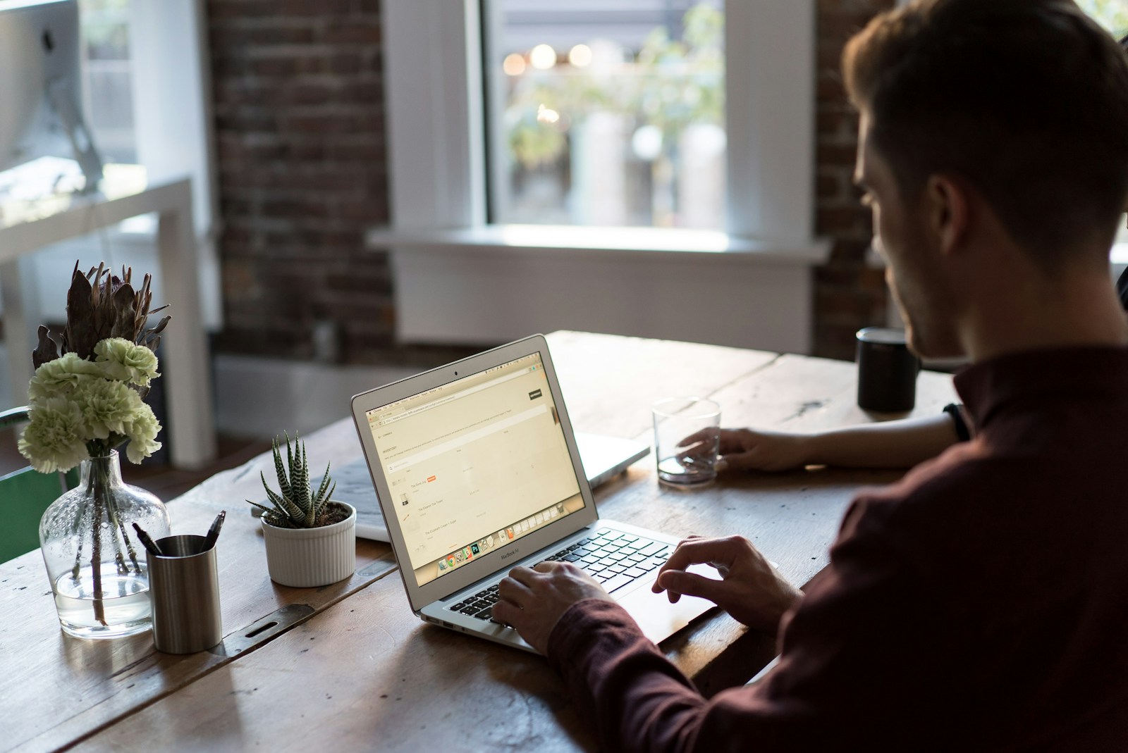 A man in a burgundy shirt types on a laptop showing an admin page at a wooden table with flowers and a succulent