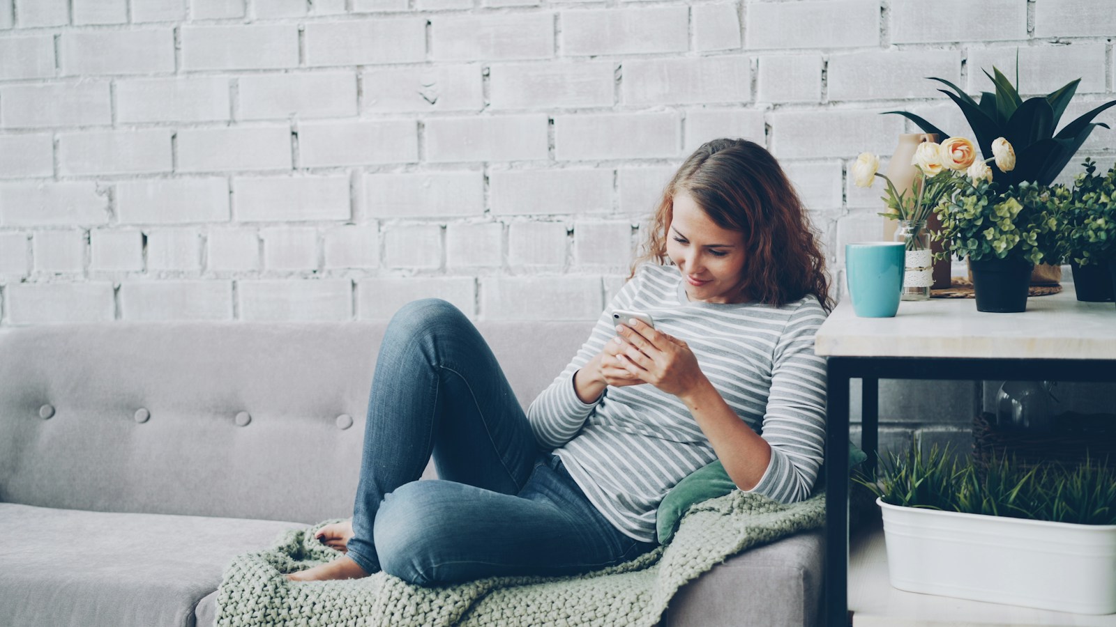 A young woman with curly hair smiles at her smartphone while lying back on a grey sofa beside a table with plants