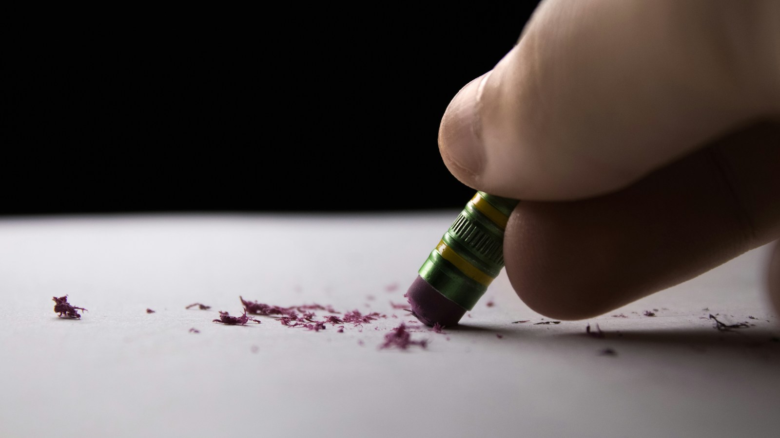 Close-up of a hand rubbing a pencil's eraser across white paper, leaving purple eraser crumbs scattered on the page