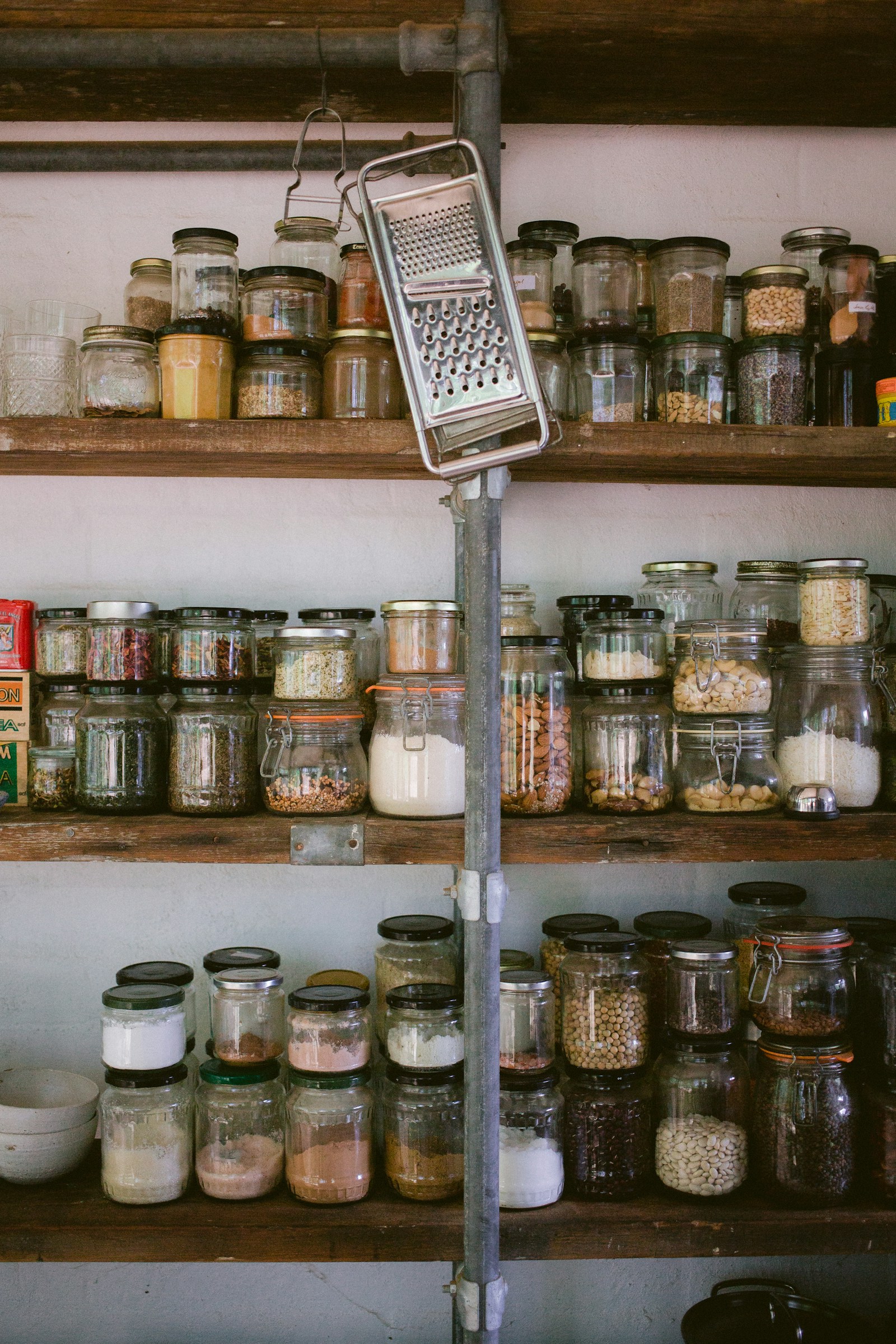Rustic wooden pantry shelves crowded with glass jars of nuts, beans, flour and spices, with a metal grater hanging in front
