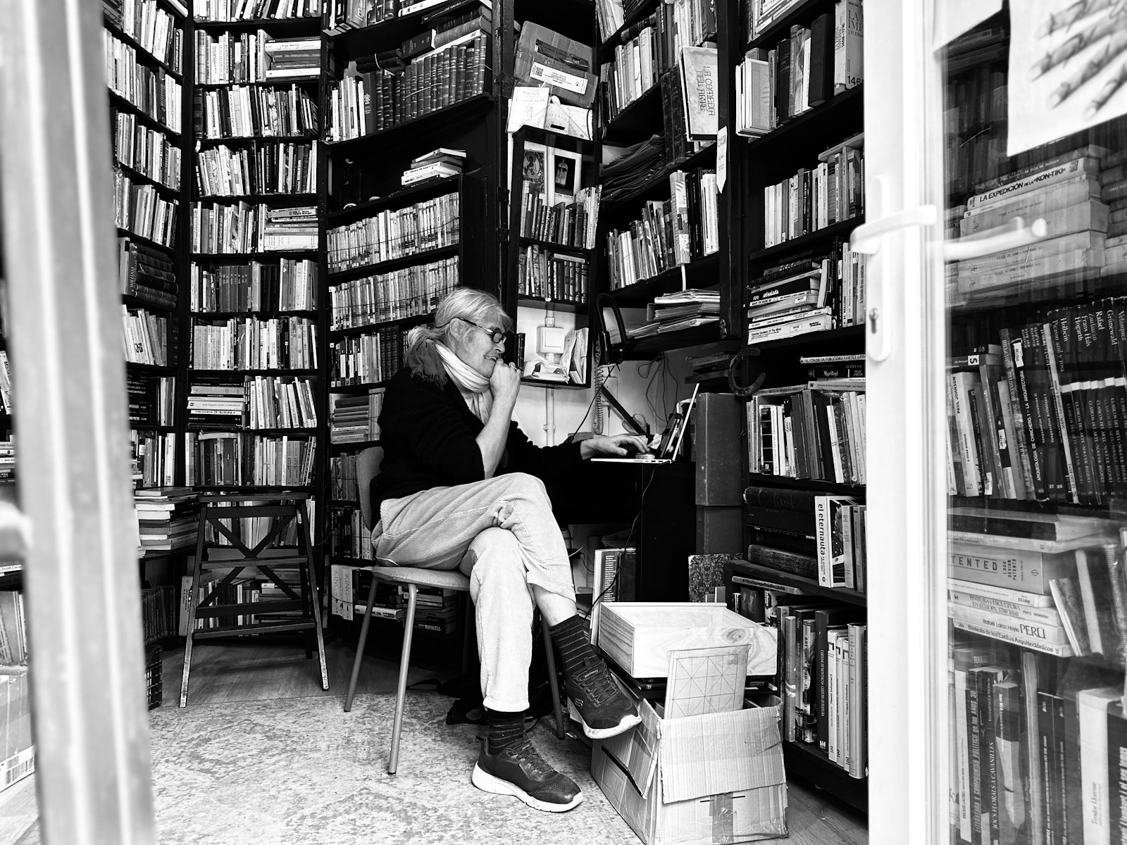 A person with glasses types on a laptop in a small bookshop lined floor to ceiling with books