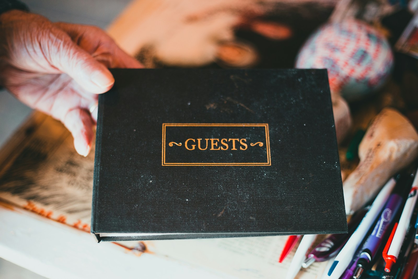 A hand holds a black guest book with GUESTS printed in gold letters above a table of pens and papers